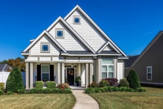 The front view of a new construction cottage craftsman style white house with a triple pitched roof with a sidewalk, landscaping and curb appeal.